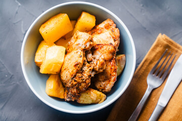 Chicken thighs baked with rustic potatoes with ju-lie sauce on a blue plate on a dark, gray background and a napkin and cutlery.