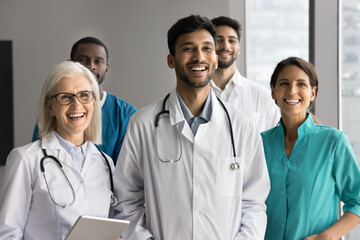 Five happy multiethnic, younger and older healthcare workers in uniforms posing for camera standing together in clinic office. Portrait of professional occupation team members, medical care and career