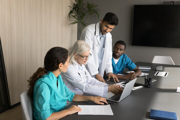 Experienced female physician and three younger colleagues, using laptop in modern meeting room, reviewing patient information, discussing treatment plans, or sharing knowledge. Teamwork, mentorship