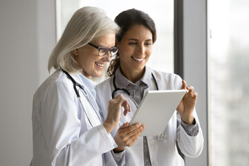 Two mature and young smiling female doctors reviewing new app or information on digital tablet, younger GP sharing details with older colleague. Mentorship, teamwork, consultation, knowledge-sharing