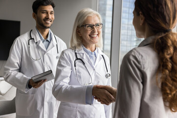 Fototapeta premium Mature female physician and intern greeting clinic patient, shaking hands, welcoming, start consultation, finishing visit in clinic, give professional medical care and help. Healthcare, communication