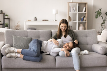 Obraz premium Happy mother rest on comfortable sofa in living room, lying on teenager daughters laps, communicate, smile, enjoy warm relationship, friendship, spend weekend time together. Motherhood, understanding
