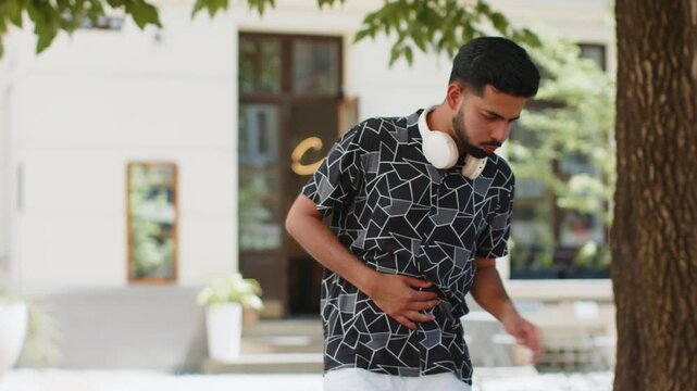 Indian man feeling sudden strong abdominal stomach liver kidney ache gastritis problem outdoors. Hispanic guy having symptom poisoning diarrhea indigestion peptic ulcer pancreatitis on city street.