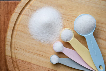 Top view of white granulated sugar in measuring spoon and pile of white granulated sugar on wooden plate for sugar level control and calorie control concept, glycemic control.