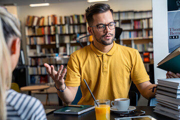 Fototapeta premium A Caucasian male and female student engage in a discussion over books in a library setting, beverages at hand, embodying a collaborative academic atmosphere.