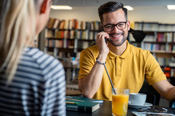 A cheerful caucasian male student converses on his phone, enjoying a cup of coffee and orange juice, seated opposite a female student in a well-stocked library setting.