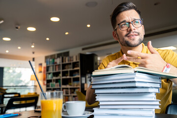 A young man leans on a stack of books, displaying contemplation, with coffee and juice nearby, in a well-lit library setting.