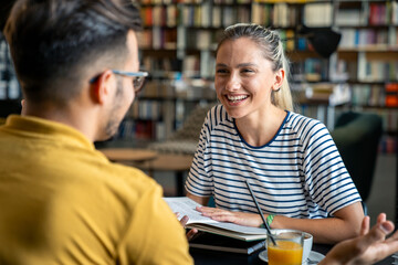 A male and female student in casual attire engage in discussion over books at a library table, with coffee and orange juice nearby.