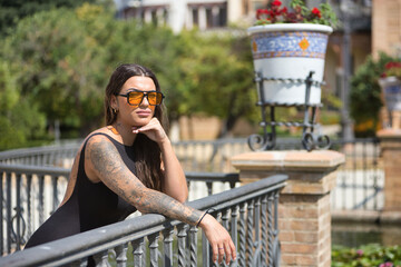 Young woman, brunette, beautiful, very tattooed, with black overalls and glasses with orange...