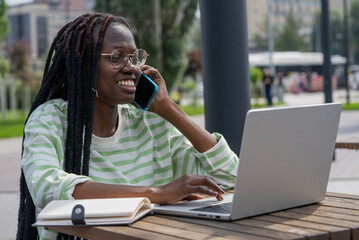 Smiling African American woman multitasking with a laptop and phone while seated at an outdoor...