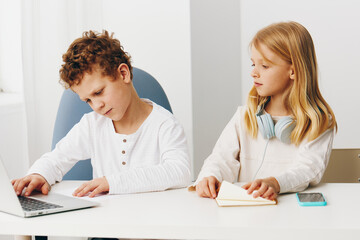 Obraz premium Happy Caucasian brother and sister sitting at home in the living room, immersed in their elearning activities The children are using laptops, tablets, and headphones, fully engaged in online education