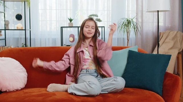 Trendy cheerful positive girl child in casual clothes having fun dancing, moving to rhythm. Teen child kid dancing emotionally rejoicing in success at home in living room sitting on sofa. Lifestyles.