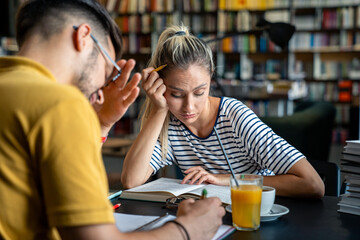 A male and female student in casual attire discuss educational content over coffee and orange juice among rows of books.