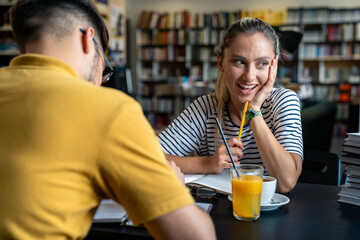A Caucasian male and female student engage in a focused study session at a library table, surrounded by books, with coffee and orange juice on hand.