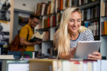 A cheerful woman engages with a tablet, while a man peruses bookshelves in a well-stocked library, both students immersed in their academic pursuits.