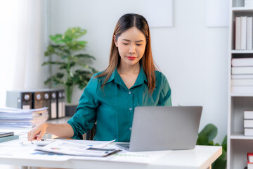 Focused on Success: A determined businesswoman diligently works at her desk, analyzing paperwork and utilizing a laptop in a bright, modern office environment. Her focused gaze and organized workspace