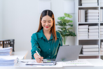 Focused and Efficient: A young Asian businesswoman, dressed in a vibrant green blouse, sits at her desk, immersed in work on a laptop and tablet, showcasing professionalism and dedication.  