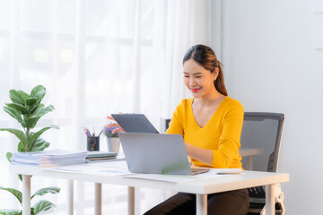 Smiling Woman Working on Laptop and Tablet:  A professional woman in a yellow shirt smiles as she works on a laptop and tablet at her desk in a bright, modern office. The image conveys a sense of prod