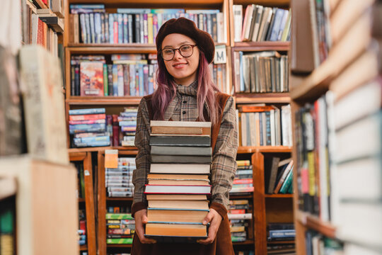 Portrait of a beautiful smart 20s hipster high school girl pupil college student teenager in glasses holding stack of books in university library or bookshop prepare for exam doing home work