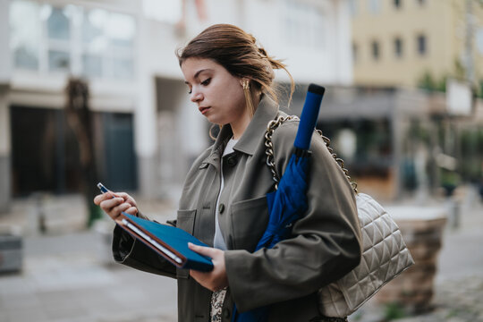 Young woman holding an umbrella and checking her notebook while walking on a city street, displaying focus and diligence.