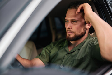 Pensive man sitting in a car looking thoughtful with a contemplative expression, dressed in a green...