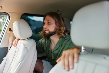 Young man with long hair sitting in a car, looking outside, wearing a casual green shirt in a light colored interior