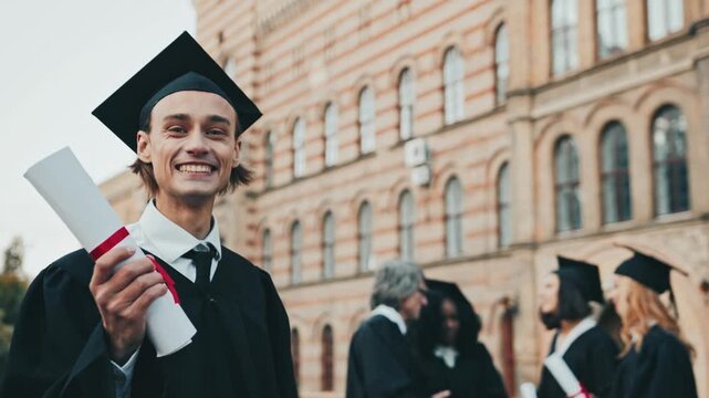 Handsome Caucasian guy wearing hat and gown on graduation day. Holding diploma rolled up into tube. Happy about finishing education in colleague or university. Happy graduates standing in background.