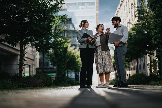 Business colleagues enjoying a friendly conversation outside an office building surrounded by greenery on a sunny day.