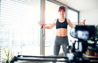 Fototapeta premium Redhead woman's home fitness routine with resistance band. Home workout on gym mat in living room, sporty woman wearing sportswear.
