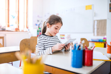 Portrait of cute schoolgirl sitting at desk using tablet during class, learning with technology....