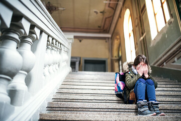 Portrait of bullied girl is crying on the steps in front of the school, waiting for her parents. Student girl with bag after day in elementary school.