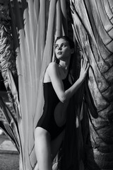 Black and white photo of a woman in a swimsuit leaning against a palm tree by the sea