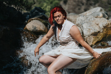 Woman in a white dress with red hair sitting by a rocky waterfall enjoying the peaceful natural surroundings.