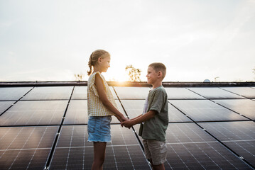 Two young siblings on roof with solar panels, looking at each other. Rooftop solar or photovoltaic system. Sustainable future for next generation.