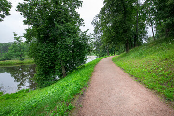 A peaceful walkway meanders beside a calm lake, framed by vibrant green trees and soft grass, inviting a stroll in the refreshing morning atmosphere.