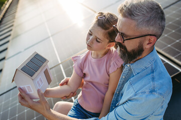 Daughter and father on roof with solar panels, holding model of house with rooftop photovoltaic...