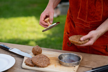 Man in apron preparing meat for hamburgers, standing outdoors. Using burger patty press to shape groud meat.