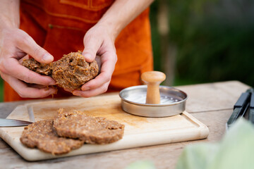 Man in apron preparing meat for hamburgers, standing outdoors. Using burger patty press to shape groud meat.