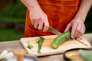 Close up of man holding sharp knife, peeling cucumber. Preparing vegetables for an outdoor barbecue.