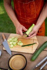 Close up of man holding sharp knife, peeling cucumber. Preparing vegetables for an outdoor barbecue.