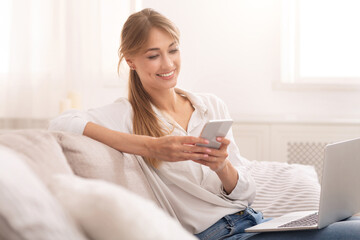 Smiling Woman Using Mobile Phone And Laptop Relaxing Sitting On Couch At Home. Selective Focus