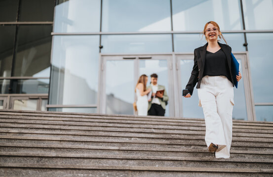 Confident businesswoman descending stairs of a modern office building, holding documents and phone, with colleagues interacting in the background.