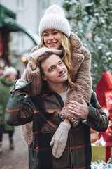 Beautiful young loving couple embracing, kissing, enjoying winter vacation, cold snowy day on a street city Christmas market outdoors. Warm clothes, knitted sweater, mittens. Cozy atmosphere