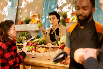 Young woman with an apron working at farmers market booth, offering fresh organic fruits and veggies. Female farmer grasping an apple and looking at camera while standing behind harvest fair stall.