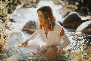 A young woman in a white dress peacefully sits by a river, enjoying the serene atmosphere of nature.