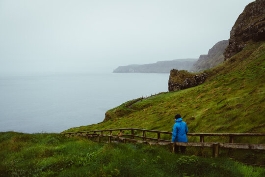 Frau mit M&uuml;tze und blauer Regenjacke auf dem Wanderweg and der K&uuml;ste der Causeway Coastal Route zur H&auml;ngebr&uuml;cke von Carrick-a-Rede in Nordirland