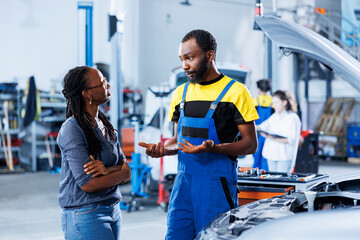BIPOC repairman helping customer with car checkup in auto repair shop. Garage worker looking over automobile parts with woman, mending her broken vehicle fuel tank during inspection