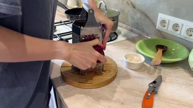 Close up of hands grating beetroot on a box grater on a wooden cutting board. Various kitchen tools and fresh ingredients are visible on the countertop
