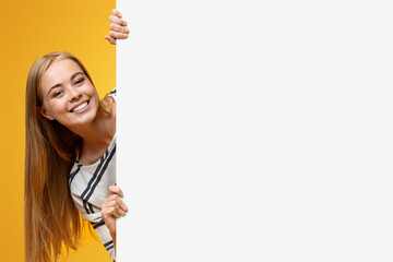 Cute teenage girl looking out of white blank placard for your text or product, yellow background