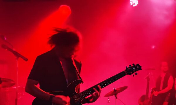 A guitarist performs on stage in front of a crowd, his hands seen strumming an electric guitar under the red light of the venue.